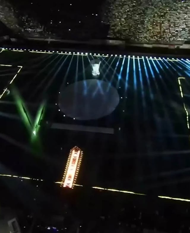 Vista aérea de un estadio de fútbol captada desde un dron, con el campo y las gradas iluminados por mini píxeles y túneles de tubos LED, que crean un espectáculo de luz y color