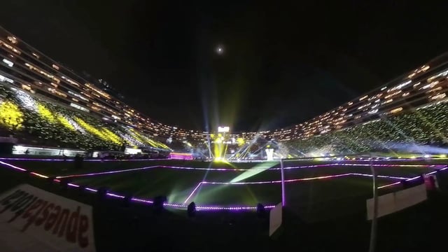 Vista desde el exterior de un estadio de fútbol con un túnel triangular de tubos LED en la entrada, que invita a los visitantes a pasar por él y disfrutar de una experiencia sensorial de luz y sonido