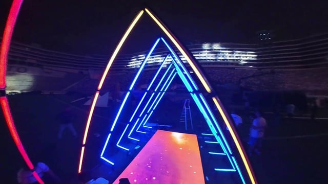 Vista desde el exterior de un túnel de píxeles LED por el que pasa un jugador de fútbol con su hija en brazos, sonriendo y saludando a la cámara, con los píxeles LED creando un fondo colorido y llamativo