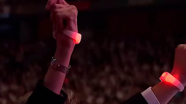 Vista desde la entrada del estadio de una mujer colocándose un brazalete luminoso al ingresar al evento en el estadio, con el brazalete mostrando el logo del evento y el color del equipo que apoya
